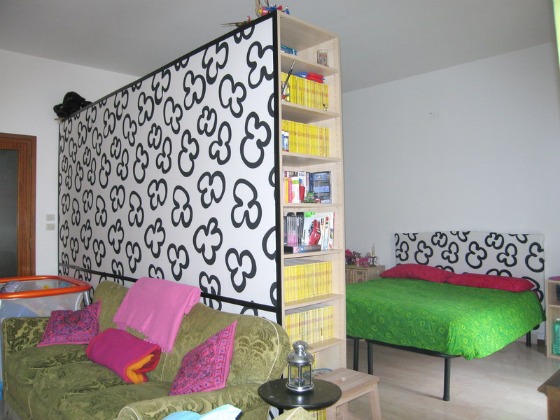 A living space and bedroom divided by a large IKEA hack bookshelf in the center with black and white designs