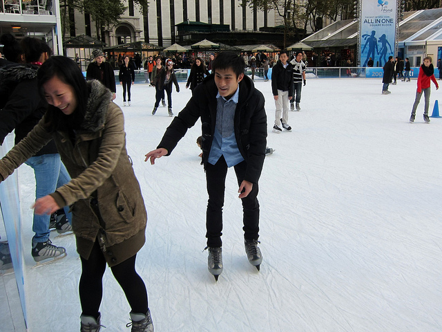Two smiling ice skaters grab for edge of Bryant Park's ice skating rink before they fall onto the ice