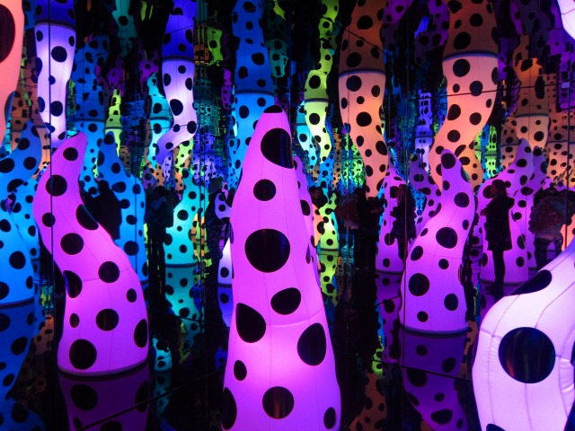 Inside Yayoi Kusama Infinity Room, called Love is calling, all the lights are off and hanging from the ceiling and bursting from the floor are brightly colored and black spotted blow up fixtures that lit up the room