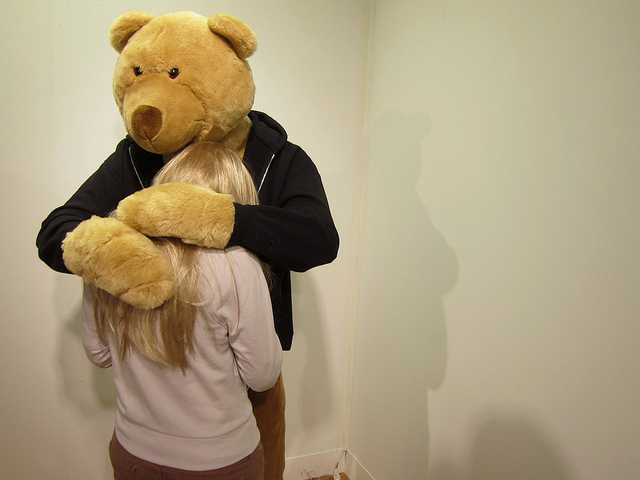A man dressed up like a teddy bear wearing a black hoodie hugs a young blonde girl