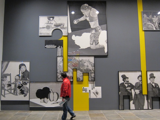 Man in a red jacket walks by a large grey and yellow wall filled with artwork by Karl Haendel at the Whitney Biennial