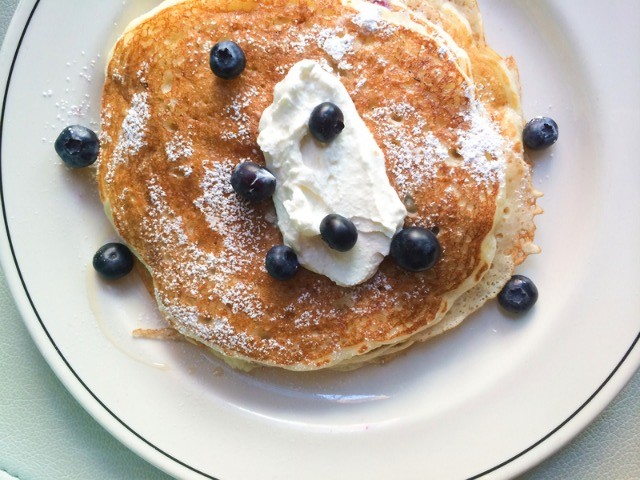 A white plate with a stack of large pancakes topped with blueberries and whipped cream