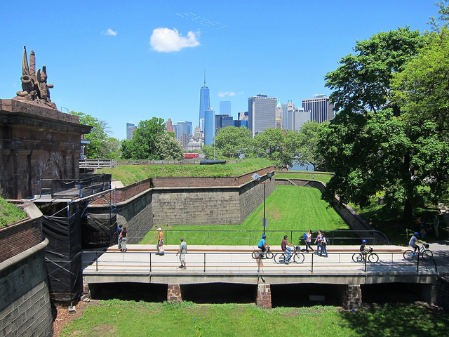 Up high on hill looking down at the new Fort Jay on Governors Island with people walking and riding bikes and the NYC skyline in the background.