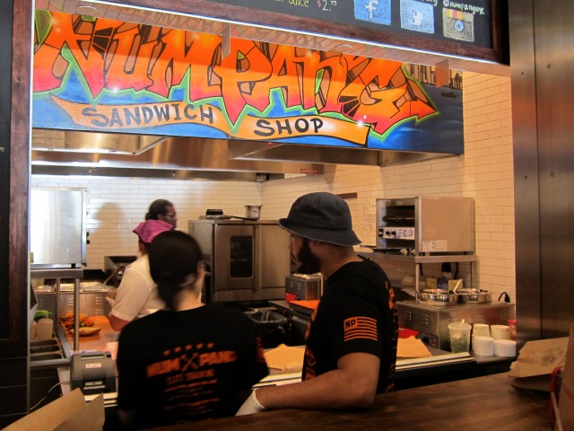 A man and a women stand in Hudson Eats at the Num Pang window to order sandwiches.