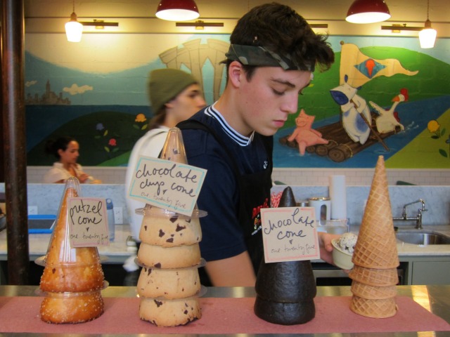 An employer scoops ice cream behind the counter of Ample Hills Creamery which displays assorted housemade cones.