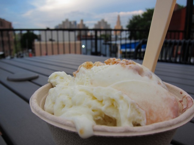 Ice cream in a cup sits on top of a picnic table with the Manhattan skyline in the background.=
