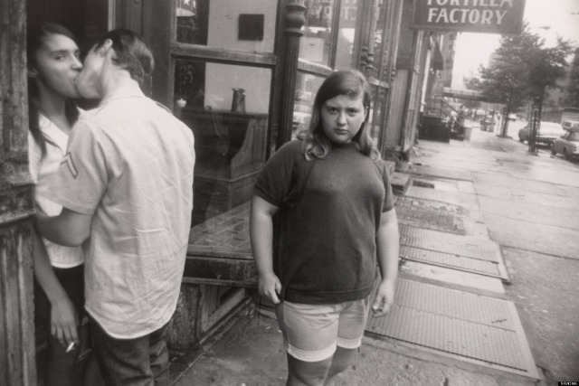 A photograph taken by Garry Winogrand of a couple kissing on the street with a young girl looking at the camera behind them.