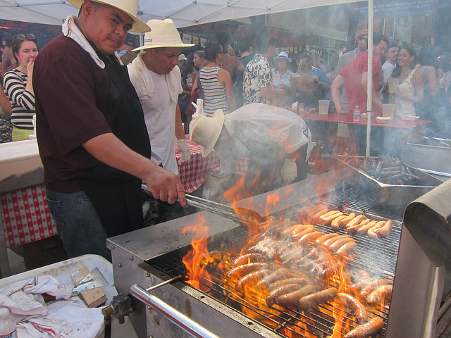 Man tending a fiery grill with hot dogs, sausage, and other meat at the Bastille Day Food Festival