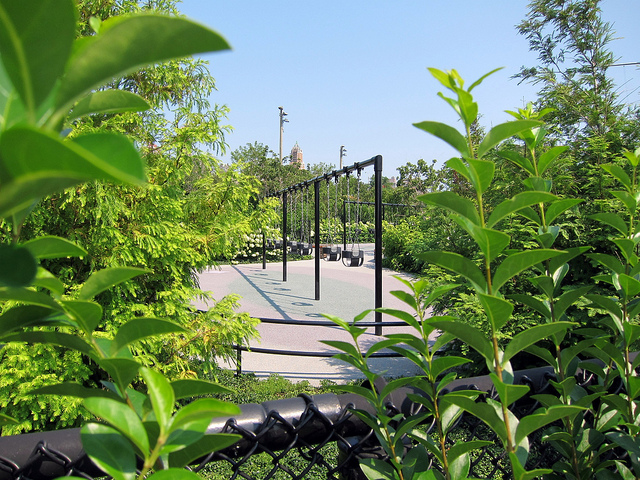 Through the bushes at the Brooklyn Bridge Park stands a large set of swing sets over a sanding clearing.