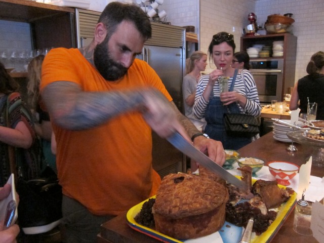A man in an orange shirt uses a large knife to cut the crust on Miss Lilly's game pie.