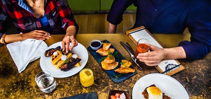 A woman in a flannel long sleeved shirt and a man in a blue button down sitting at a distressed table enjoying brunch