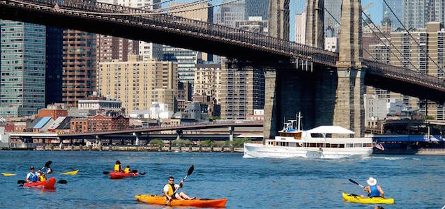 A group of kayakers under the Brooklyn Bridge in New York City on a sunny day.