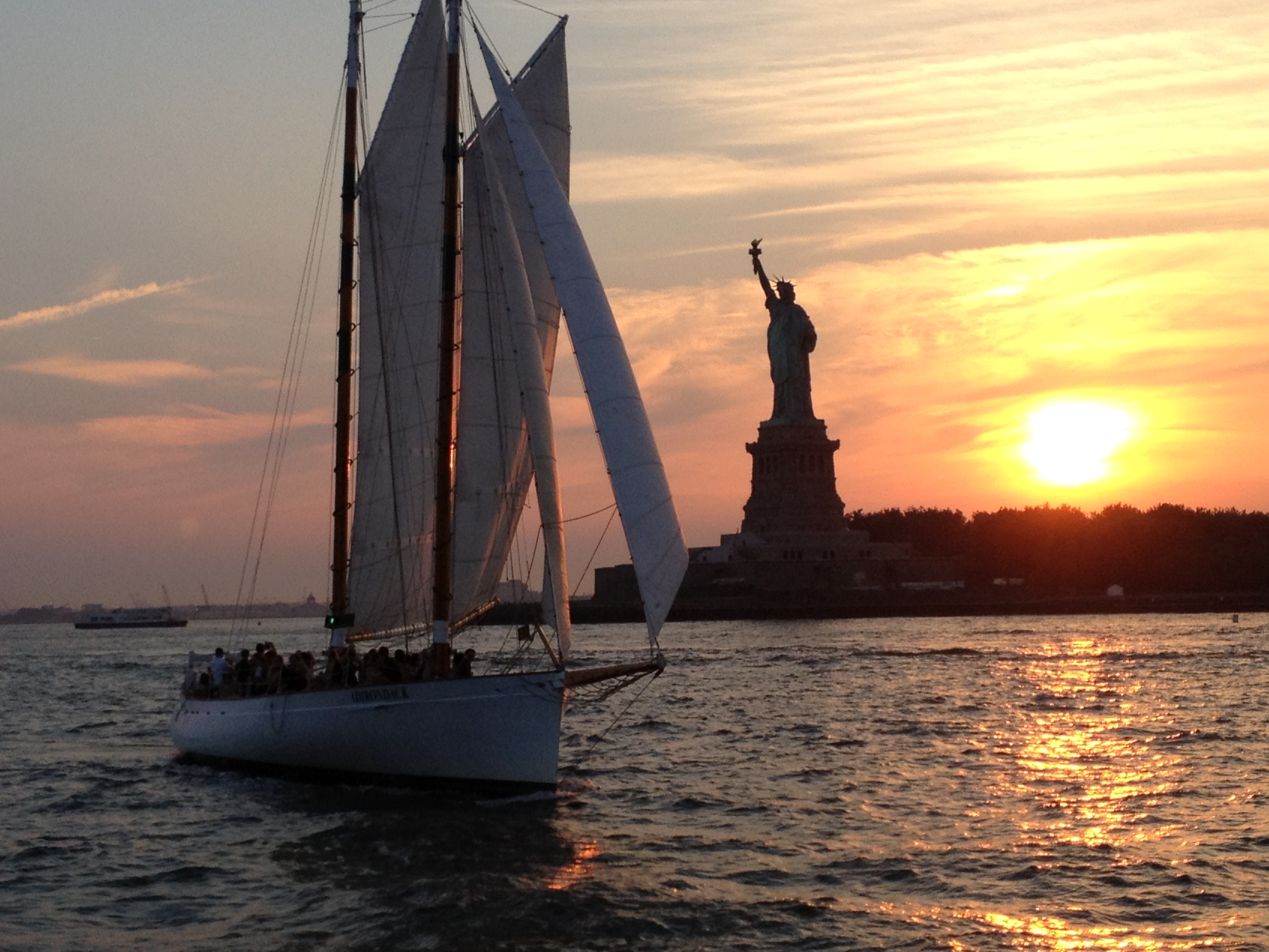 A group of individuals sailing by the Statue of Liberty into a summer sunset.
