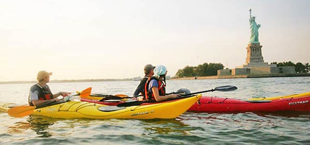 A group of kayakers around the Statue of Liberty in New York City at sunset.