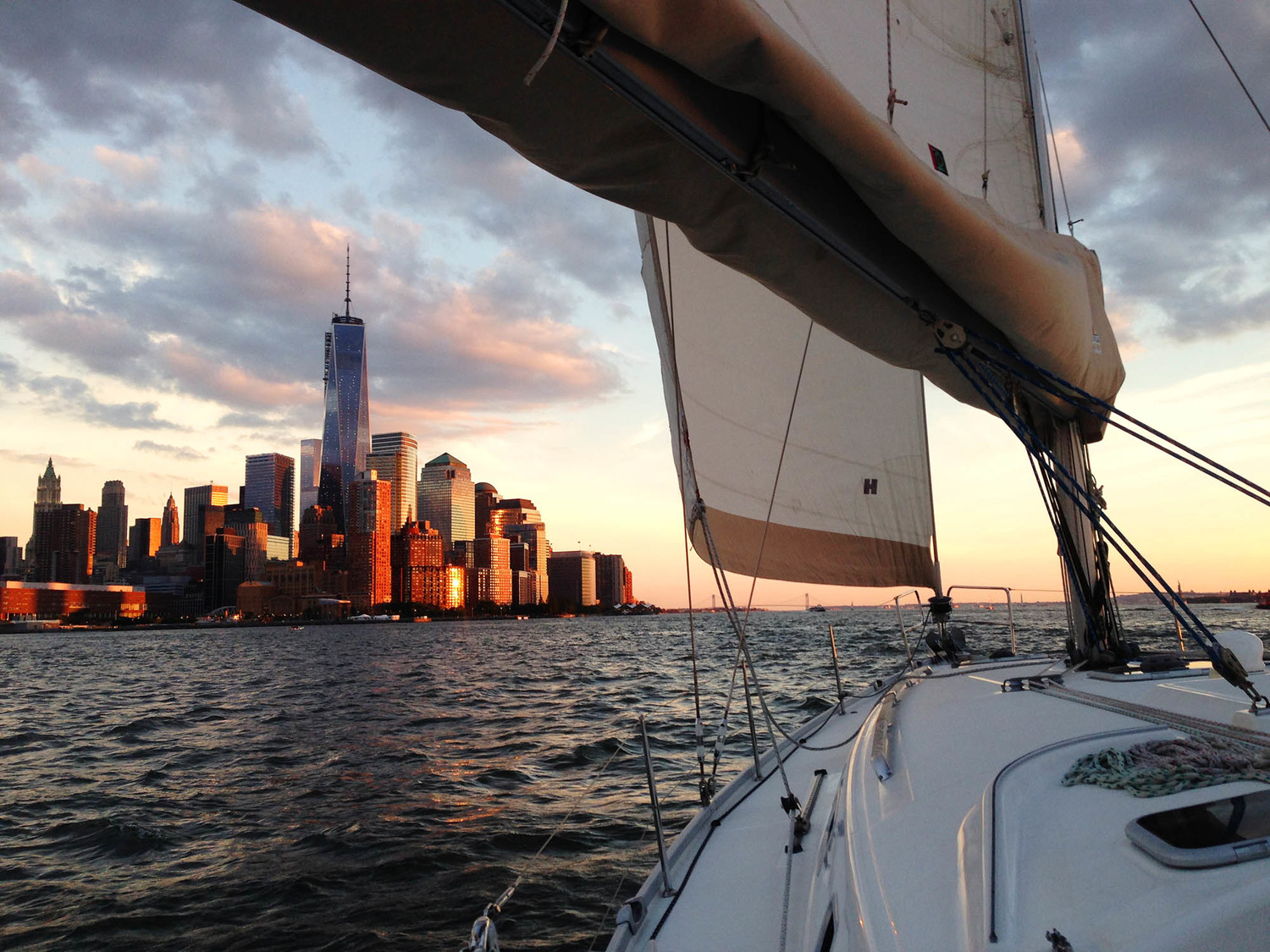 A starboard view of a sailboat with the New York City skyline, at dusk to the left of them from a sailors point of view 