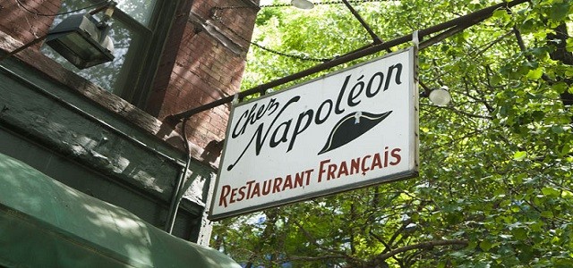 The storefront sign of French bistro Chez Napoleon hanging from the brick building in front of green leaves and trees in the sunshine.