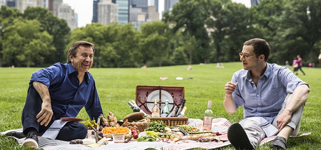 Daniel Boulud sitting outside on a sunny day in a park with green grass having a picnic with a man with a prepared picnic basket and bottle of wine.