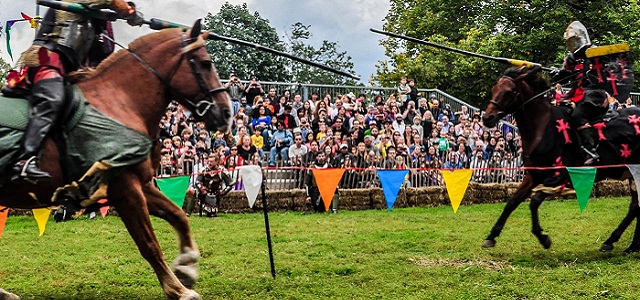 Two men on horseback at the Fort Tryon Medieval Festival practicing jousting in front of a large audience in the stands outside on a sunny day.