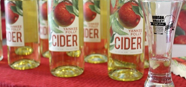 Glass bottles of cider with apple son the label and a nearby tall glass on a table with a red covering at the Hudson Valley Wine & Food Festival.