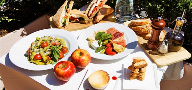 An outdoor picnic on a wooden table with glasses of water, pasta salad, paninis, fresh bread and olive oil with two apples in the sunlight outside.