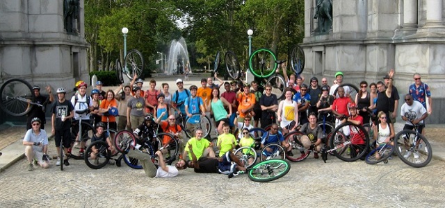 A group of cyclists with their unicycles posing for a picture outdoors in front of a water fountain and stone structure.