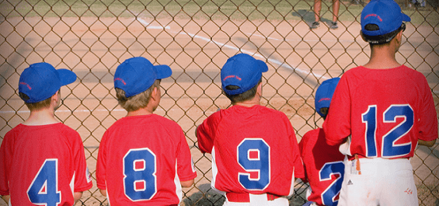 A group of 4 young boys in matching red and blue uniforms watching a baseball game being played from behind a fence on a sunny day.