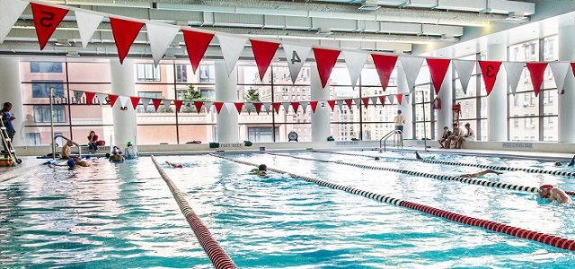 An indoor pool surrounded by large windows with sunlight coming through as swimmers pass under red and white strings of flags crossing the blue pool water.