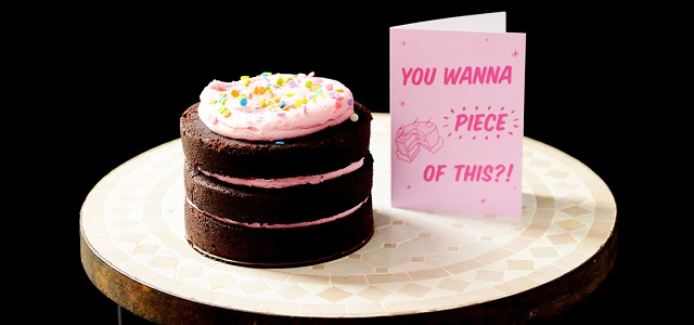 A vegan three layer chocolate cake with pink icing and rainbow sprinkles next to a pink card that says "You wanna piece of this?!" on a cream tiled table.