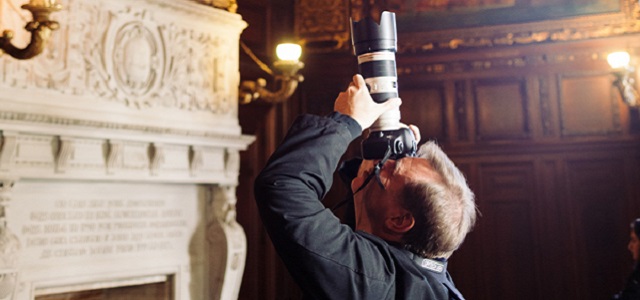 A photographer points his camera for a shot with dim lighting in the background of brown wooden walls and a white stone fireplace.