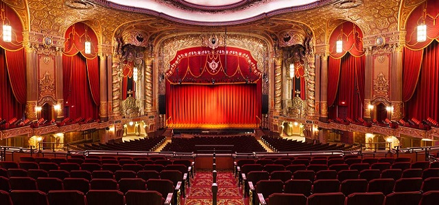 An interior view of a golden theater with bronzed wall and bright red drapes and curtains with deep maroon seating.