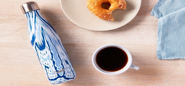 A navy, light blue, and white swirl design on a Swell water bottle on a light brown wooden table near a black cop of coffee and a bitten bagel on a plate.