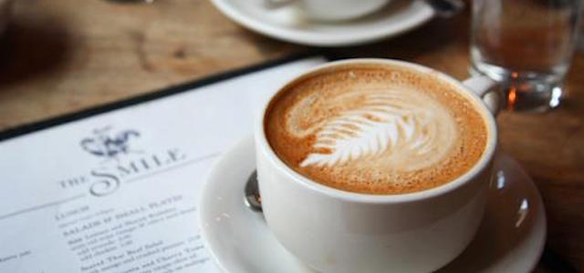 A cup of coffee with a leaf design sitting on a brown wooden table at The Smile in NYC.