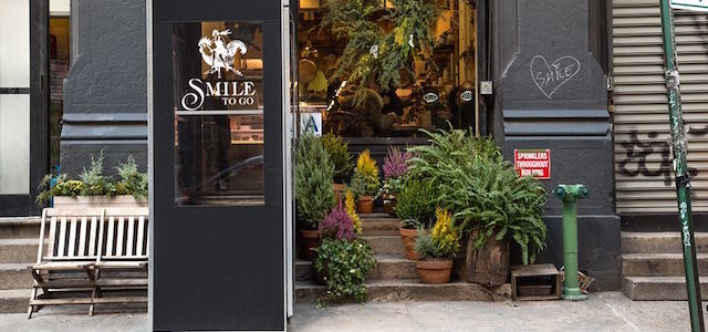 A view of The Smile's front door entrance during the day with blooming flowers and a bench.