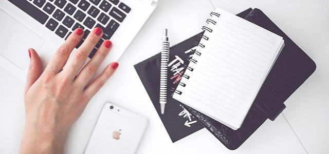 A woman is wearing bright red nail polish on her hand resting on a white desk nearby a white iPhone and linked paper notebook.