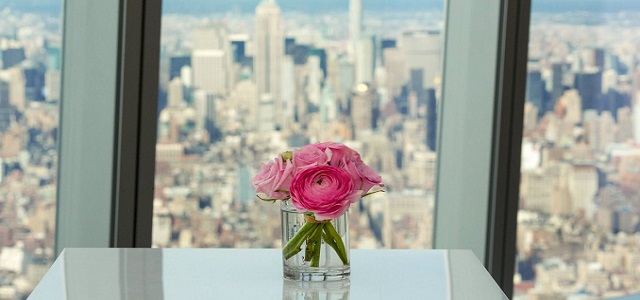 A glass with bright pink flowers in front of a clear window overlooking the New York City skyline from a skyscraper.