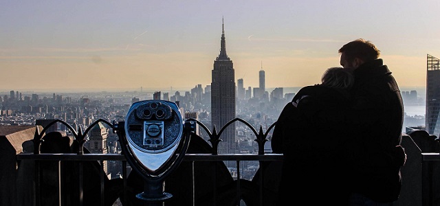 A couple hugging on top of the Empire State Building at sunset with the NYC skyline in the background.