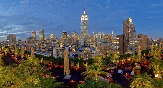 An outdoor view of 230 Fifth's outdoor patio with lights and greenery overlooking NYC skyline at  night.