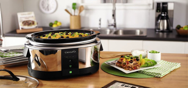 A stainless steel crock pot with fresh food sitting on a wooden kitchen counter with plates and cup nearby.