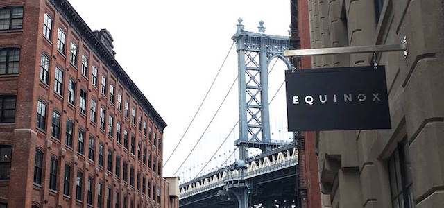 An exterior view of an Equinox location in New York City with a black sign hanging on a stone building near a bridge.