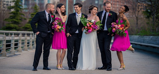 A group of men and women taking part in a big fat wedding in NYC wearing hot pink attire and black suits.