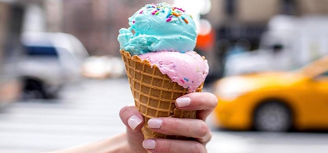 A ice cream cone with blue and pink scoops help outside near a yellow NYC taxi.