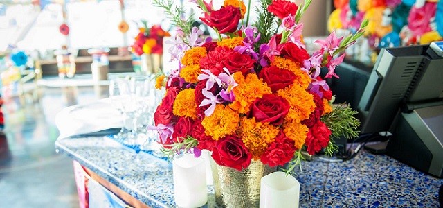 Brightly colored fresh flowers in a metallic vase sit on a blue desk in the sunlight.
