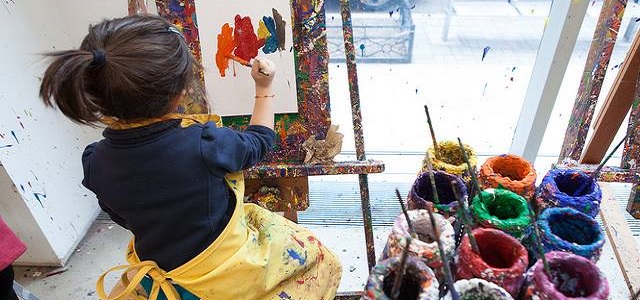 A small brunette girl wearing a yellow apron sitting in front of a large sunny window painting on an aisle. 