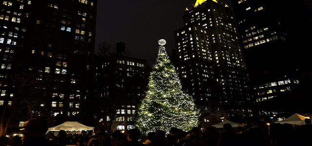 A lit Christmas tree at night in New York City with tall buildings in the background.