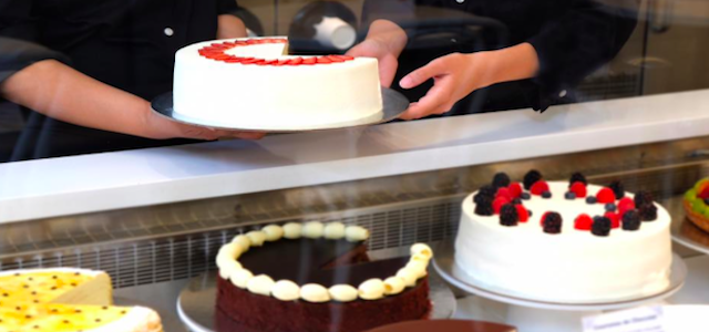 Two bakers cutting a decorated cake with white icing over a display case.