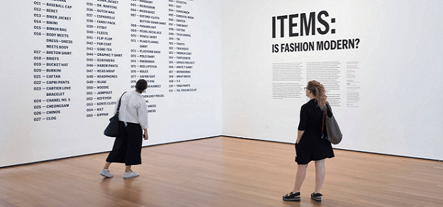 Two women standing at MoMA reading a fashion exhibit.