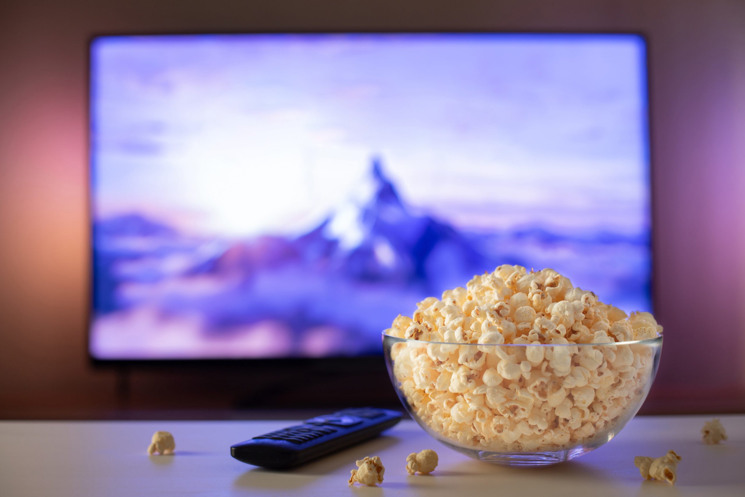 A bowl of popcorn and a clicker on a coffee table with a tv in the background