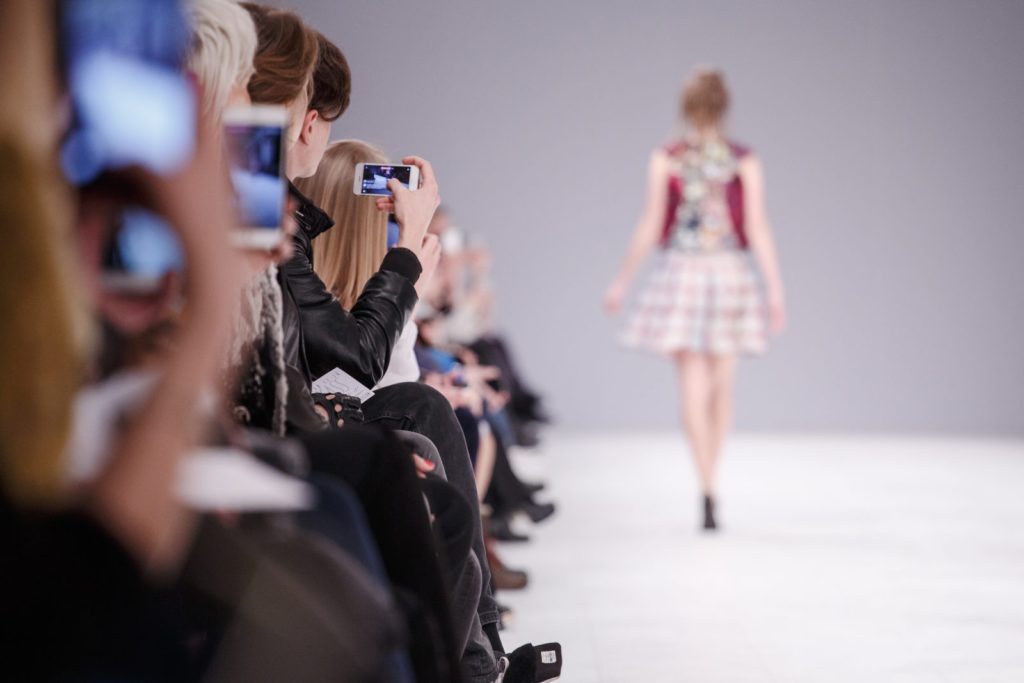 A thin fashion model wears a white plaid skirt as photographers take her picture walking down a white runway.
