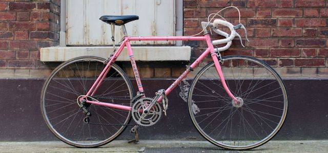 A light pink bike with a black seat leaning against a brick building.