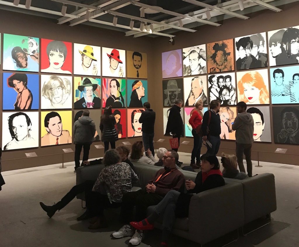 A group of adults stand and sit on couches and admire an artwork collection of famous actors and actresses in a museum.